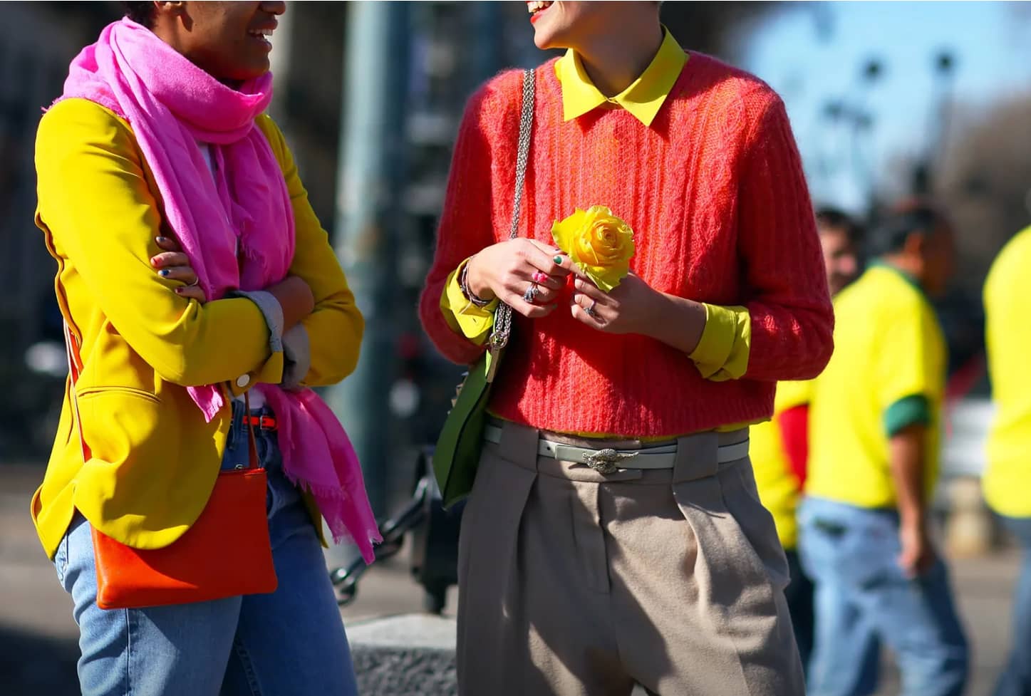 Two people on the street at Colville Spring 21.  We see their mid-frame where one wears a yellow shirt with an red jumper over it and holding a yellow flower. The second person wears a yellow jacket with a red bag and pink scarf.