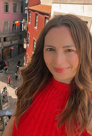 Headshot of Irish blogger Anita Kenna in red top with red lipstick and wavy brown long hair. Porto city behind her.