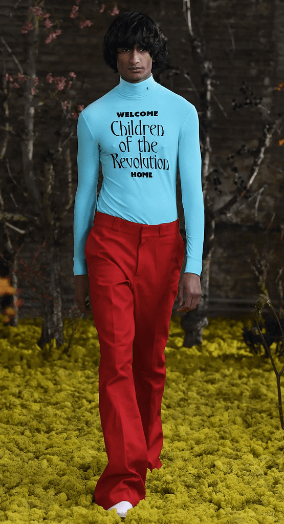 Male model on a forest style runway at Raf Simons Spring 2021 show wearing red trousers, blue top with typography on it stating "Welcome Children of the Revolution Home"