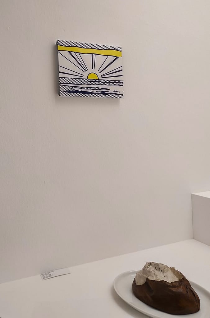 Blue, yellow, white and black small artwork of sun rising over the sea on display against a white wall in Ludwig Gallery. Work by Roy Lichtenstein, called Sunrise.