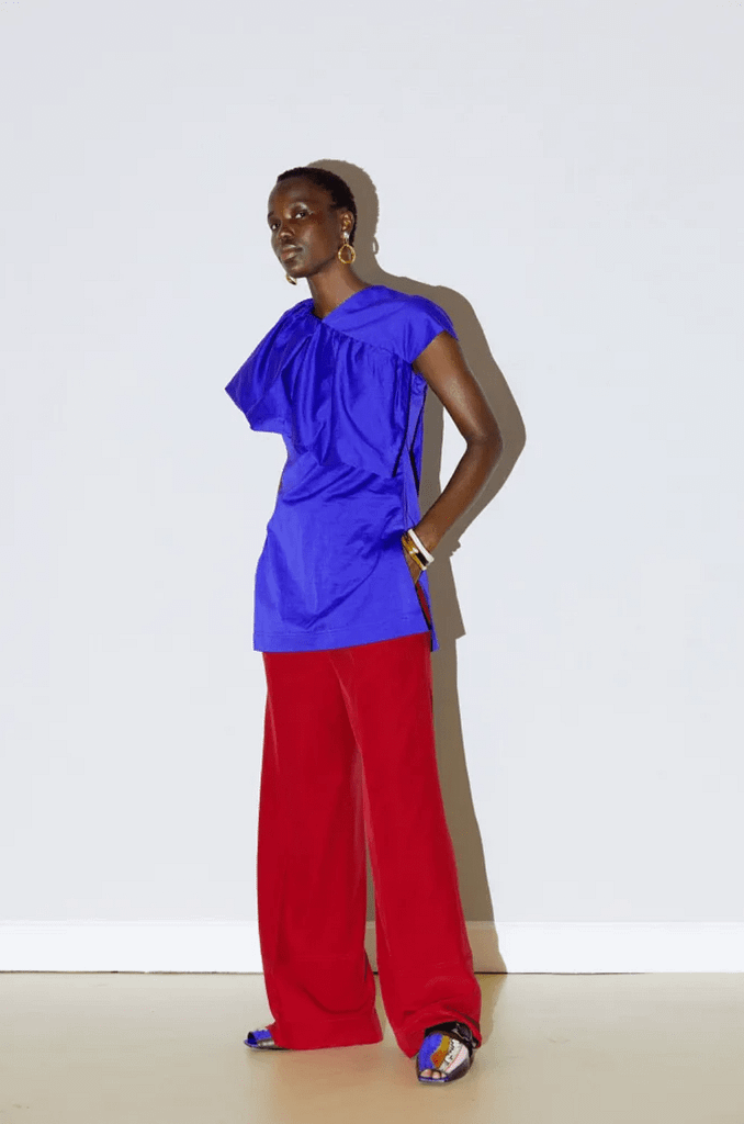 Black model standing against a white background at Colville Spring 21. Model wears blue sick top with a read trousers.