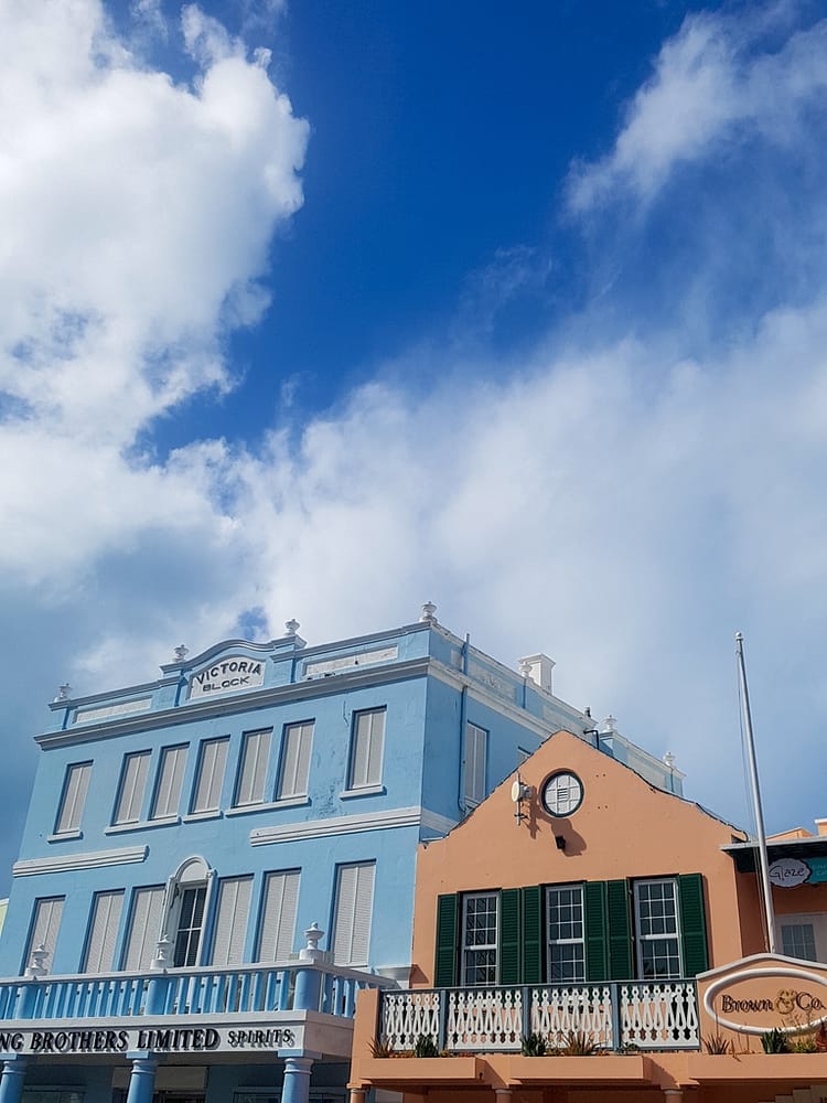 Two buildings on Front Street, Bermuda. Pastel two story blue building, with white decorative elements and window frames, stating "Victoria Block" and "Brothers Limited Spirits". Brown building with ivory shutter frames beside it. Blue sky with some clouds.