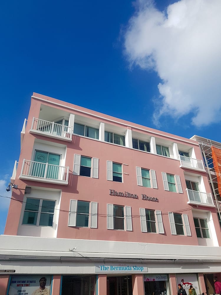 Pink three story building with white windows stating "Hamilton House" and "The Bermuda Shop" on the building. Blue sky with one cloud.
