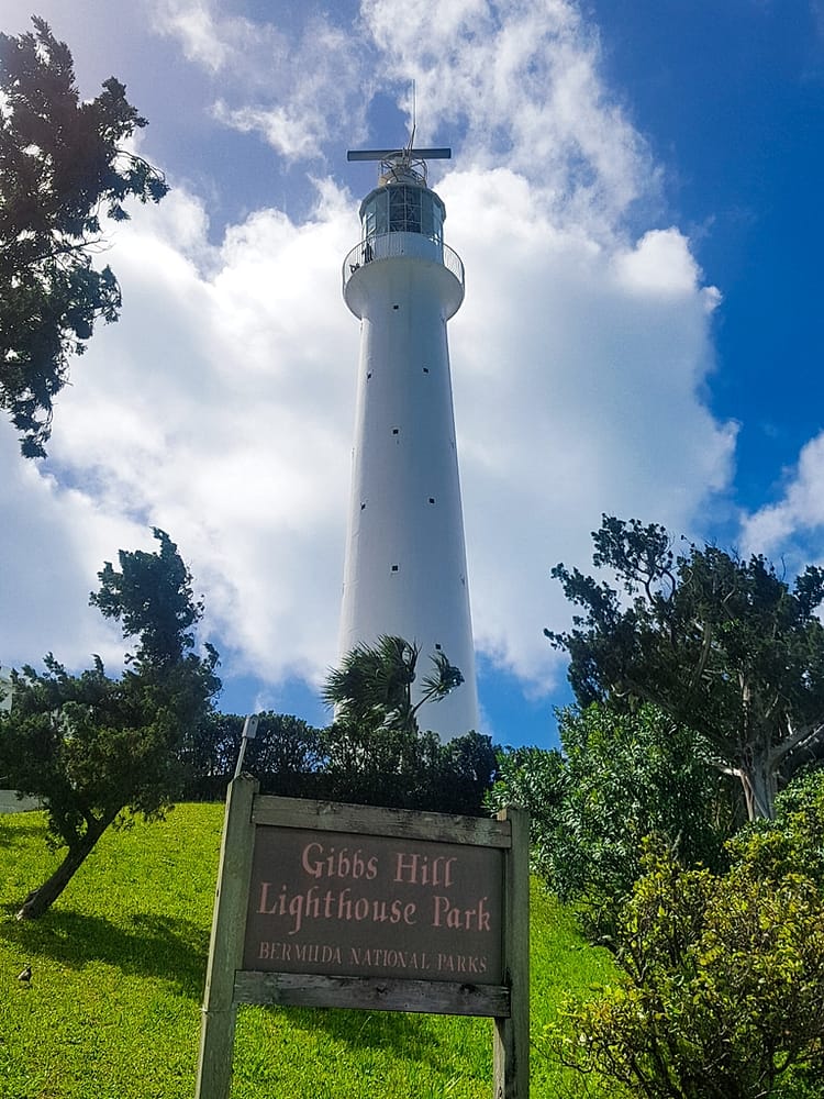 Green grass and trees with white Gibb's Hill Lighthouse. Blue sky and clouds. Sign stating "Gibbs Hill Lighthouse Park".