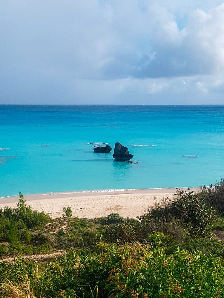 View from road overlooking greenery with a sandy beach and sea in the distance. Two rocks are emerging from the water near the shoreline at Warwick Long Beach, Bermuda.