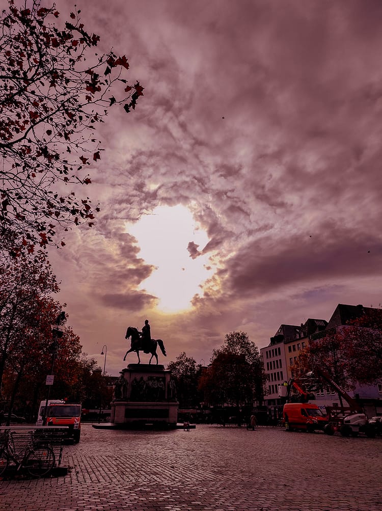 Silhouette in dark of statue of Friedrich Wilhelm III at Heumarkt Square. Tree leaves and buildings frame the image as dark shapes in the sunset too. Circle of white light from the sun shines in the sky over the statue.