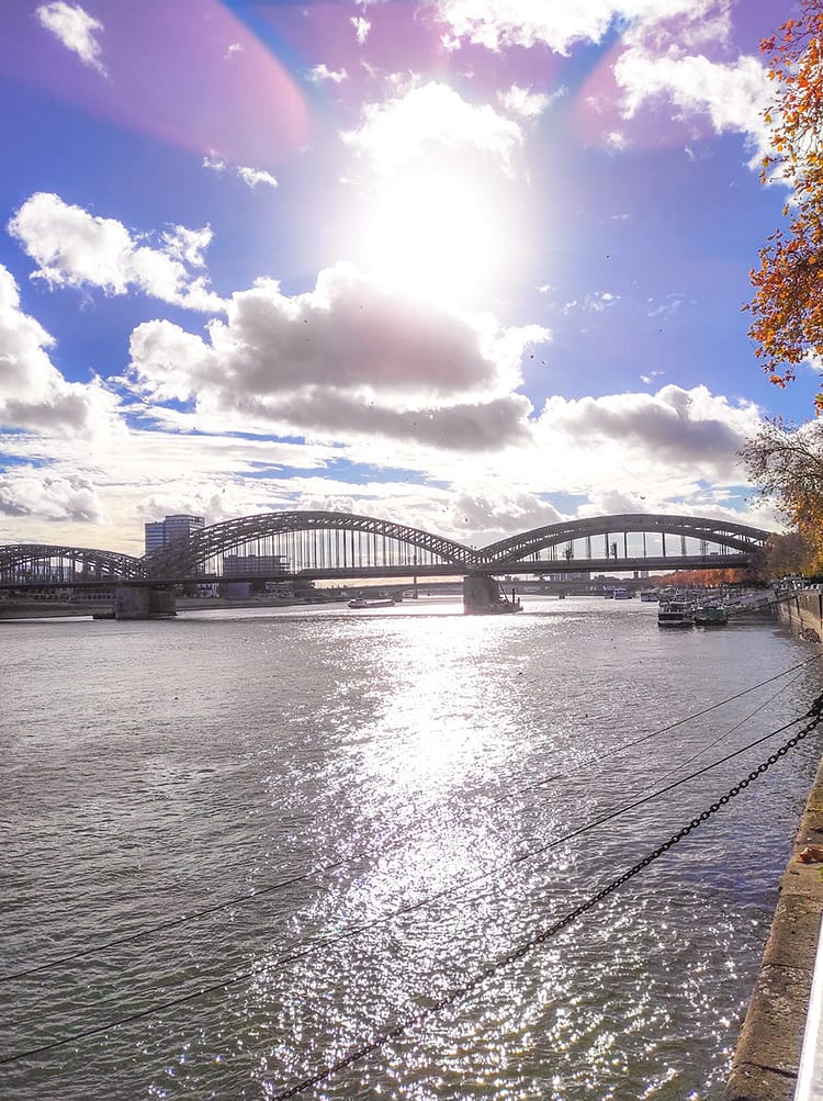 View of Rhine River and Hohenzollern Bridge in Cologne. Blue sky with some clouds and a bright sun shining over the bridge.