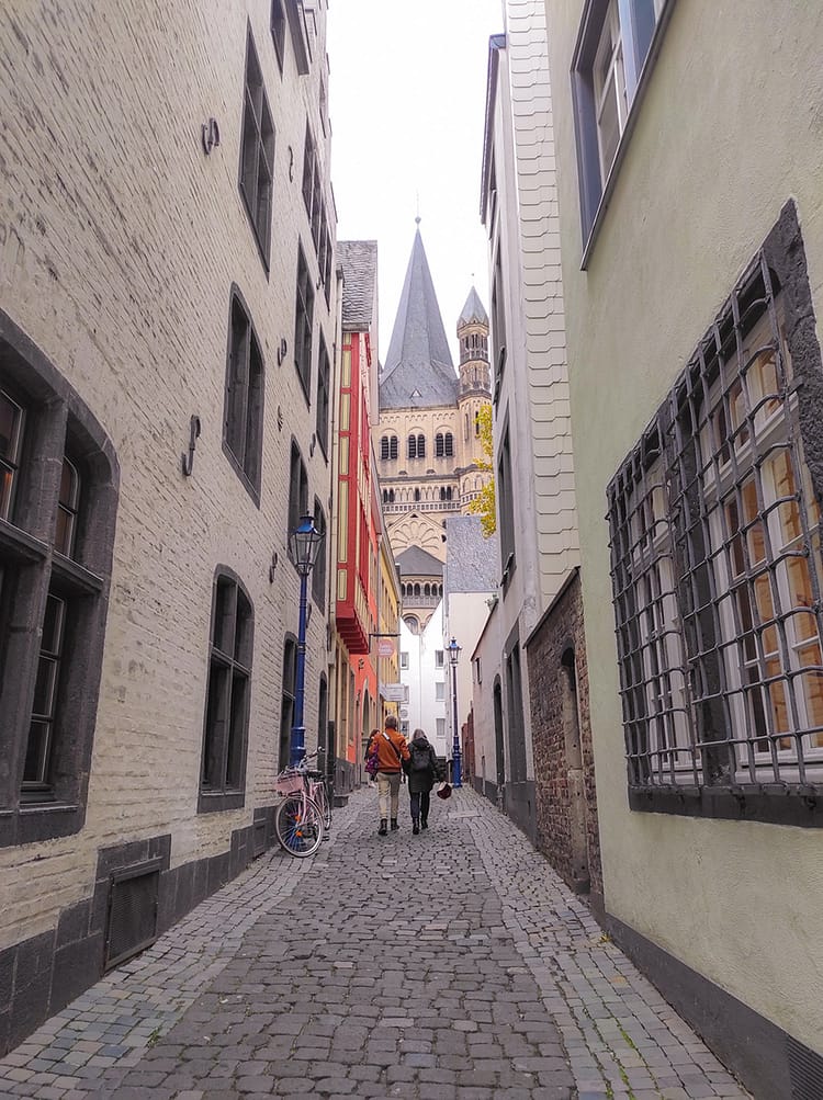 Small cobblestone street, with two people walking towards Groß St. Martin Church, in Cologne. Church is framed in the center of the image at the end of the street.