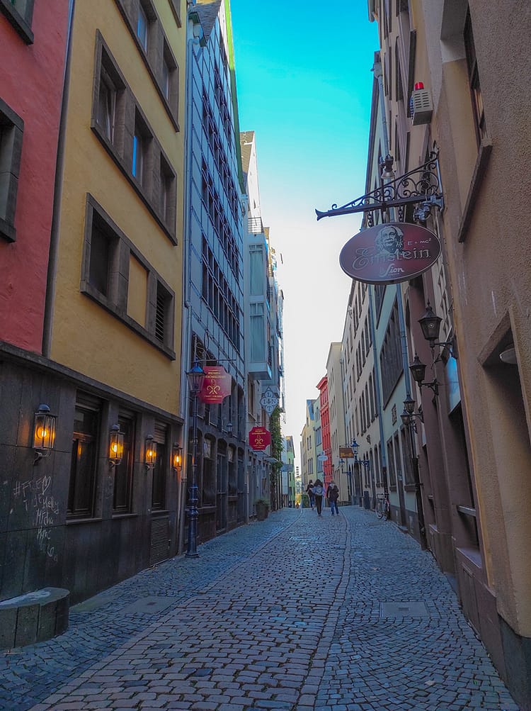 Very small dark cobble stone street, Lintgasse Street, with buildings all along it. People walking in the distance on it with blue sky overhead. Some hanging signs for bars, etc., including a sign for Einstein bar which includes an image of Einstein on the hanging sign.