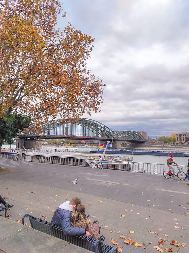 View of Hohenzollern Bridge and River Rhine in Cologne. A seat is along the walk way beside the river with a couple embracing while looking at the view. Autumn yellow and brown colored leaves on a tree in the corner of the frame over the couple.