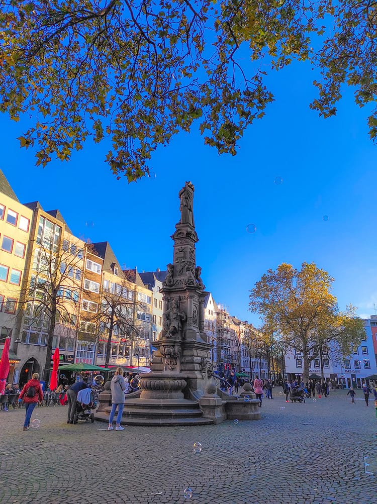 Alter Markt Square, Cologne. Cobbled square with monument of Jan von Werth-Brunnen, (which includes a fountain) found in the centre. Colorful buildings and some trees are found in the square. Bright blue sky is overhead. Children blowing bubbles by the monument.