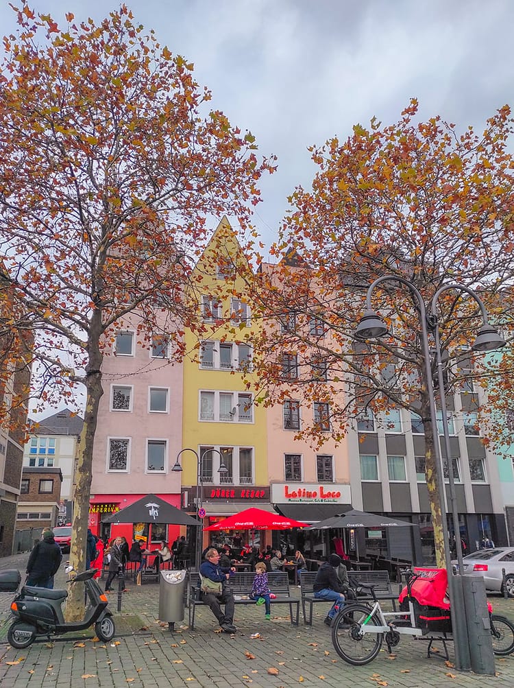 Colorful buildings along Heumarkt Square. Bikes parked at front of buildings and bench with people sitting on it sits in the middle of the frame. Trees with autumnal leaves sit along the front of the buildings at each side of the bench.