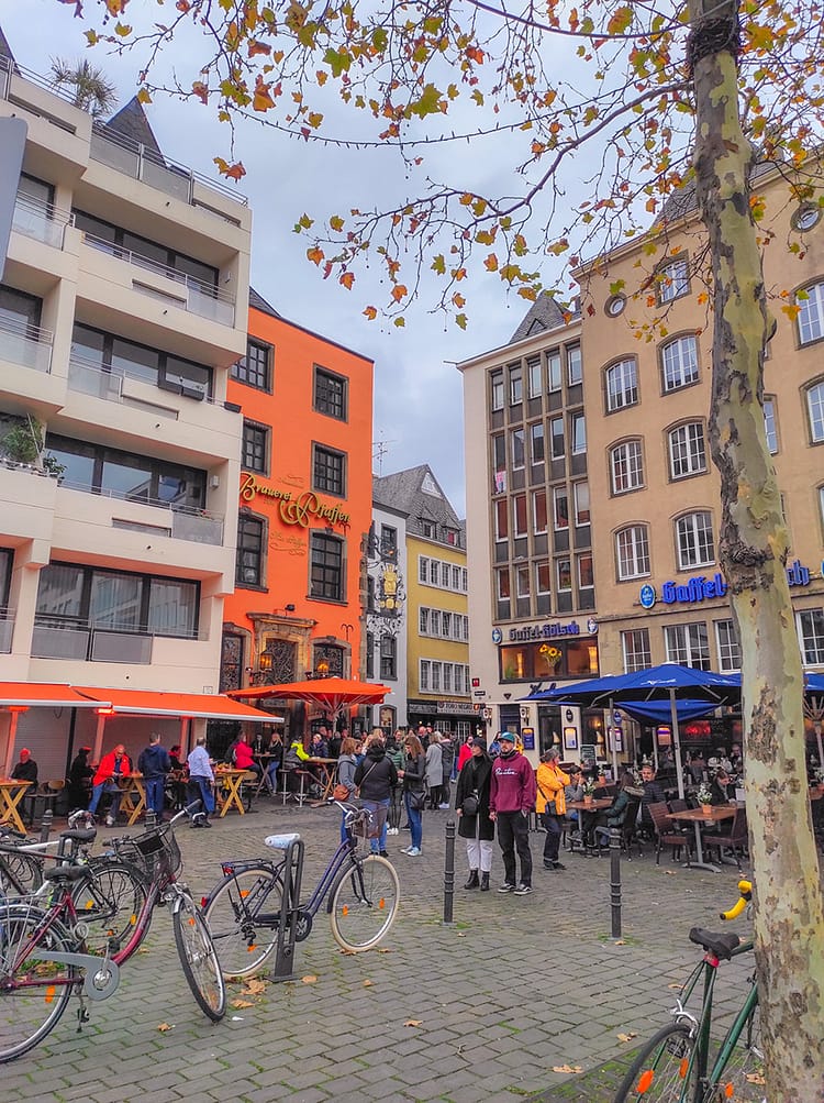 Colorful buildings at the corner of Heumarkt Square. Outdoor tables with umbrellas sit outside the buildings on top of cobblestone streets that lead off in the distance. Tree with autumnal leaves frame the top of the photograph.