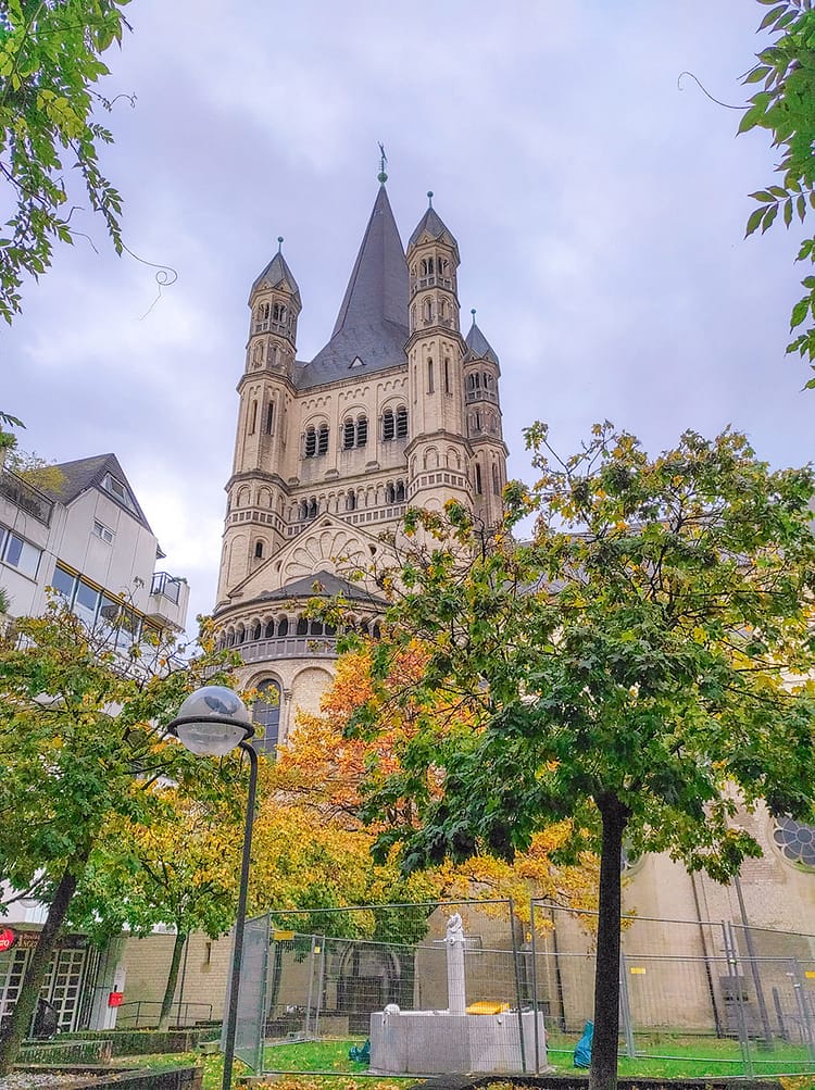 View of Groß St. Martin Church, in Cologne, surrounded by green and autumnal orange leaved trees and blue sky.