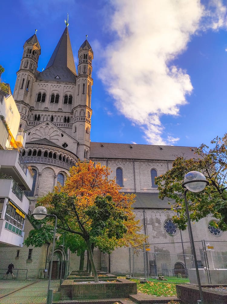 View of Groß St. Martin Church, in Cologne, surrounded by green and autumnal orange leaved trees and blue sky with one cloud overhead.