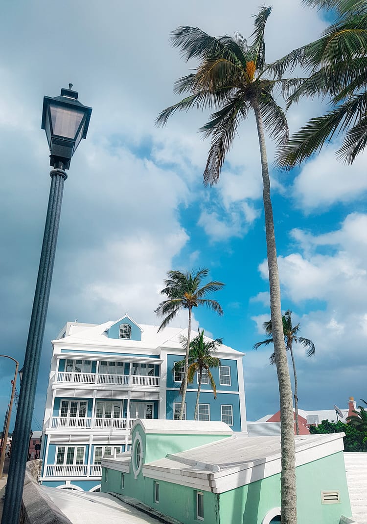 Palm trees, street lamp and large blue building with white roof, white balcony rails and window panes in Bermuda. Blue sky with clouds above the building.