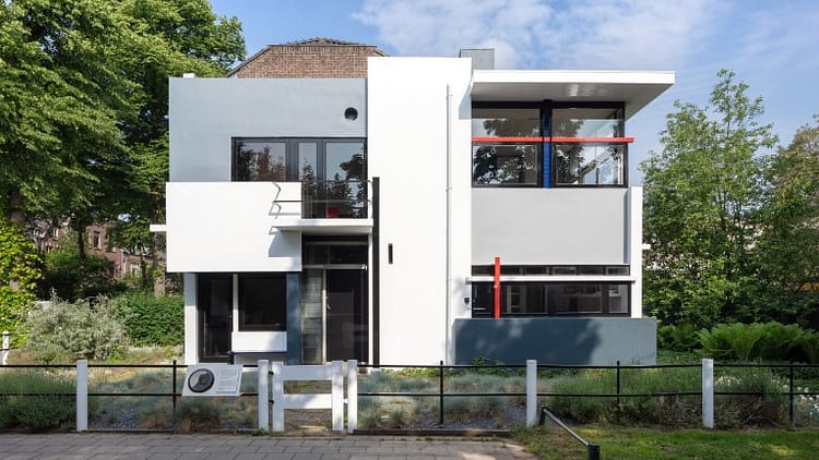 Rietveld Schroder House, De Stijl style building in white, grey and with red and blue strips across the windows, surrounded by grass, trees and with a fence in front of building