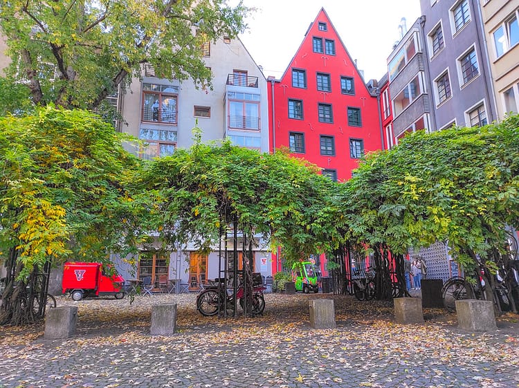View of a Cologne city square with pavement covered in fallen Autumn leaves. Green and yellow trees dot along the square with German architectural buildings in the background (including a bright red/orange building). Tünnes und Schäl monument statues lie in the corner of the square. Bicycles are parked against the trees and bright orange and green mini vehicles are parked by the buildings. 