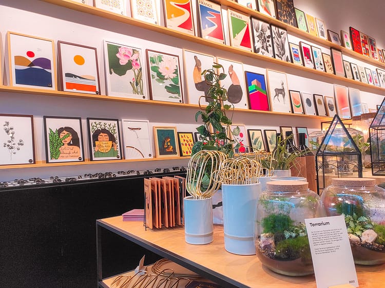 White wall with four rows of wooden shelves with colorful framed prints on them for sale in Schee, Cologne. Wooden table to the forefront of the picture, in front of the wall, with plants and home accessories on it (for sale too).