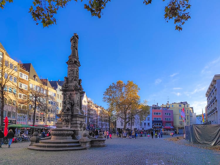 Alter Markt Square, Cologne. Cobbled square with monument of Jan von Werth-Brunnen, (which includes a fountain) found in the centre. Colorful buildings and some trees are found in the square. Bright blue sky is overhead.