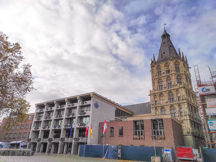 Photograph of section of City Hall (Kölner Rathaus), Cologne, with its Renaissance-style architecture. Cloudy bright sky lies above the building with some blue breaking through.