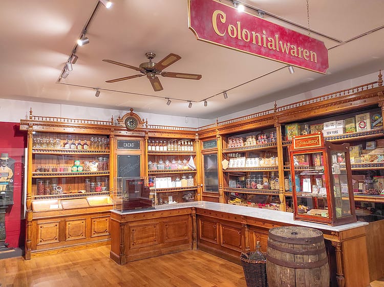 Indoor installation of the interior of a historic Colonial goods store with artifacts, such as cacoa for sale. Also, it includes a sign for Stollwerck, a German chocolate manufacturer, on a glass case.