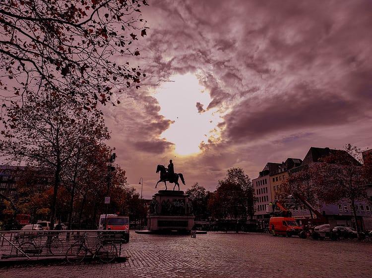 Silhouette in dark of statue of Friedrich Wilhelm III at Heumarkt Square. Tree leaves and buildings frame the image as dark shapes in the sunset too. Circle of white light from the sun shines in the sky over the statue.