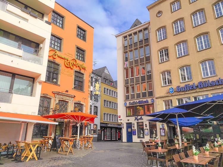 Colorful buildings at the corner of Heumarkt Square. Outdoor tables with umbrellas sit outside the buildings on top of cobblestone streets that lead off in the distance.