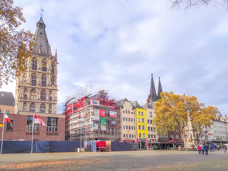 Part of City Hall, Cologne, is to the side of the frame. It has Renaissance-style architecture. Beside it sits colorful buildings on the cobbled square of Alter Markt. The top of Cologne Cathedral can be seen in the distance. Cloudy bright sky lies above the building.