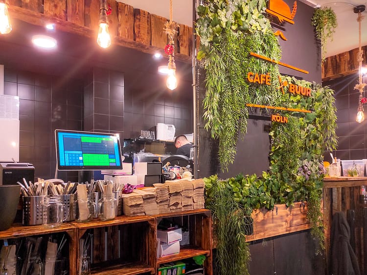 Cashier counter of Cafe Buur with wooden frame, fake greenery and an orange sign stating Cafe Buur, Koln.