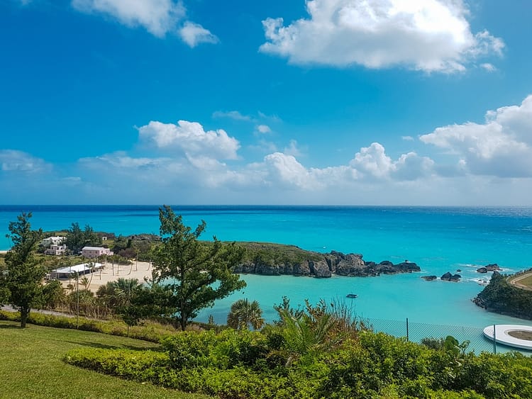 View overlooking greenery and small beach in the distance in Bermuda. Bright aqua blue sea and blue sky with some clouds. 