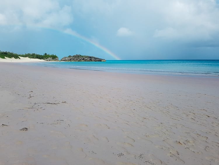 Sand, sea, some rocks and greenery in the distance. A rainbow is above the rocks at Horseshoe Bay, Bermuda.