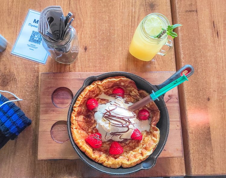 Brown wooden table with saucepan containing fluffy pancake with a chocolate syringe and melted Nutella, ice cream and strawberries. Glass jar containing freshly squeezed ginger juice and a straw on the table too.