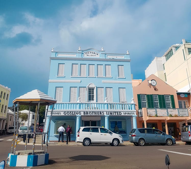 Cars on crossroads at Victoria Block, Front Street, Bermuda. Two story pastel blue Gosling Brothers building containing white decorative panels and window shutters with a brown building containing green window shutters beside it.