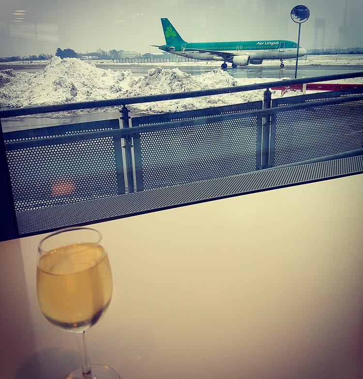 A glass of wine on a table, by the window, in Dublin Airport. Outside the window is an Aer Lingus plane on the runway surrounded by snow.