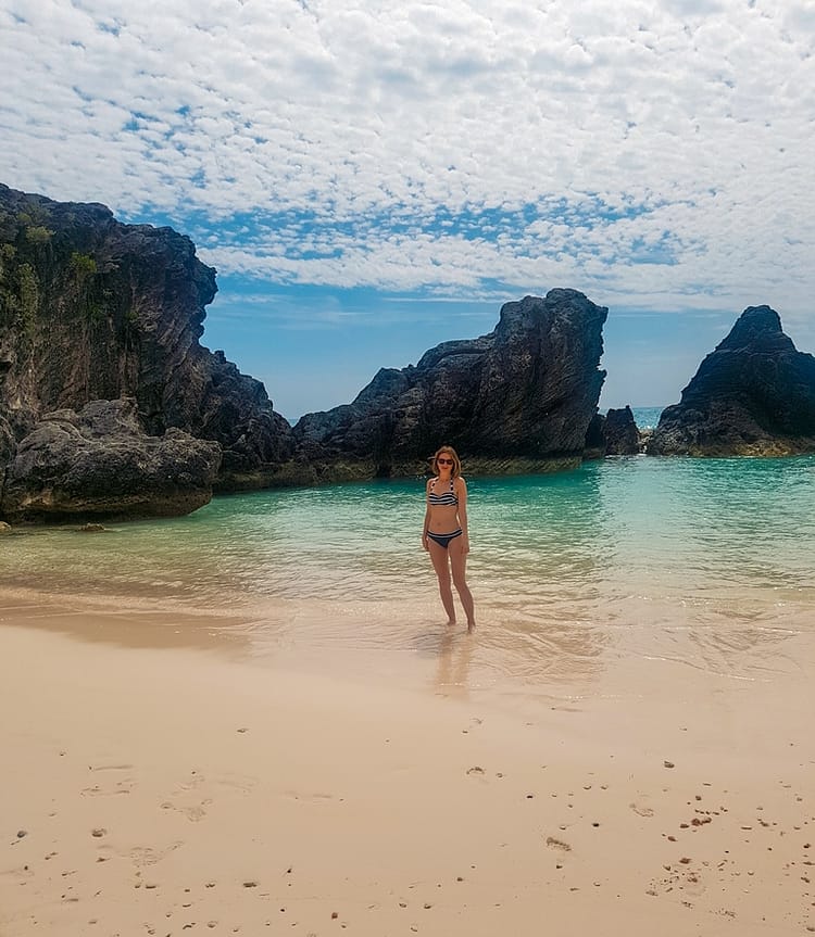 Girl in bikini standing in the sea shoreline at Jobson's Cove, Bermuda. Large dark black and grey rocks are emerged from the water behind her.