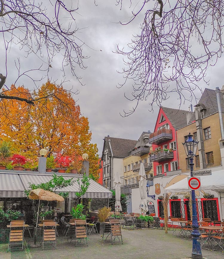 Grey sky day. Pretty square with Autumn colored leafy trees in the background and framing the top of the photo. Several restaurants with outdoor dining and covered over with umbrellas. Nice street lamp to the side of the frame which has a sign on it stating Square just off Kastellsgäßchen.