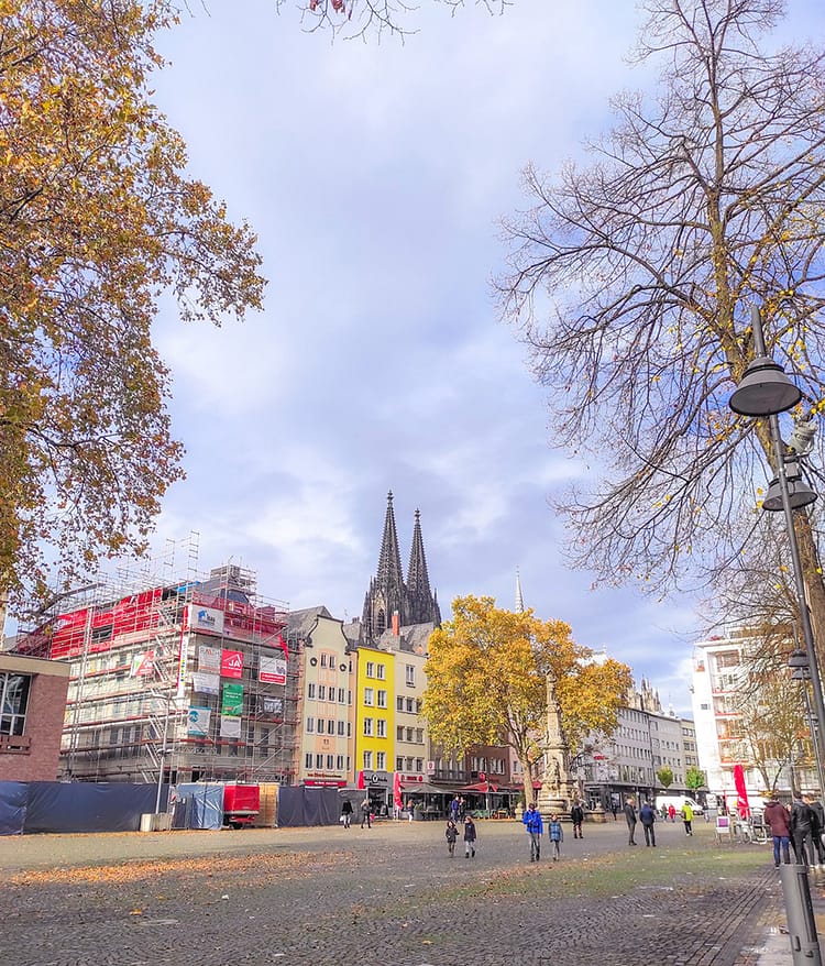 Colorful buildings on the cobbled square of Alter Markt. The top of Cologne Cathedral can be seen in the distance. Cloudy bright sky lies above the building. Autumnal leaves of trees frame both sides of the photograph.