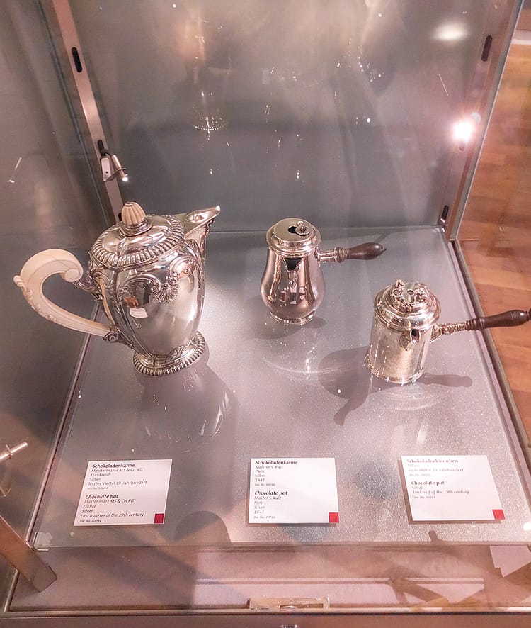 Three historic silver chocolate pots on display behind glass in Cologne Chocolate Museum.