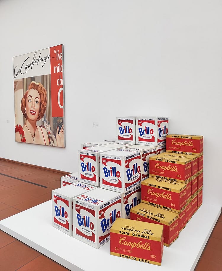 Photography of Ludwig Museum room where Andy Warhol's Brillo Boxes are on display on on a white board on the floor in gallery. Pop art work on the white wall beside the Brillo boxes too.