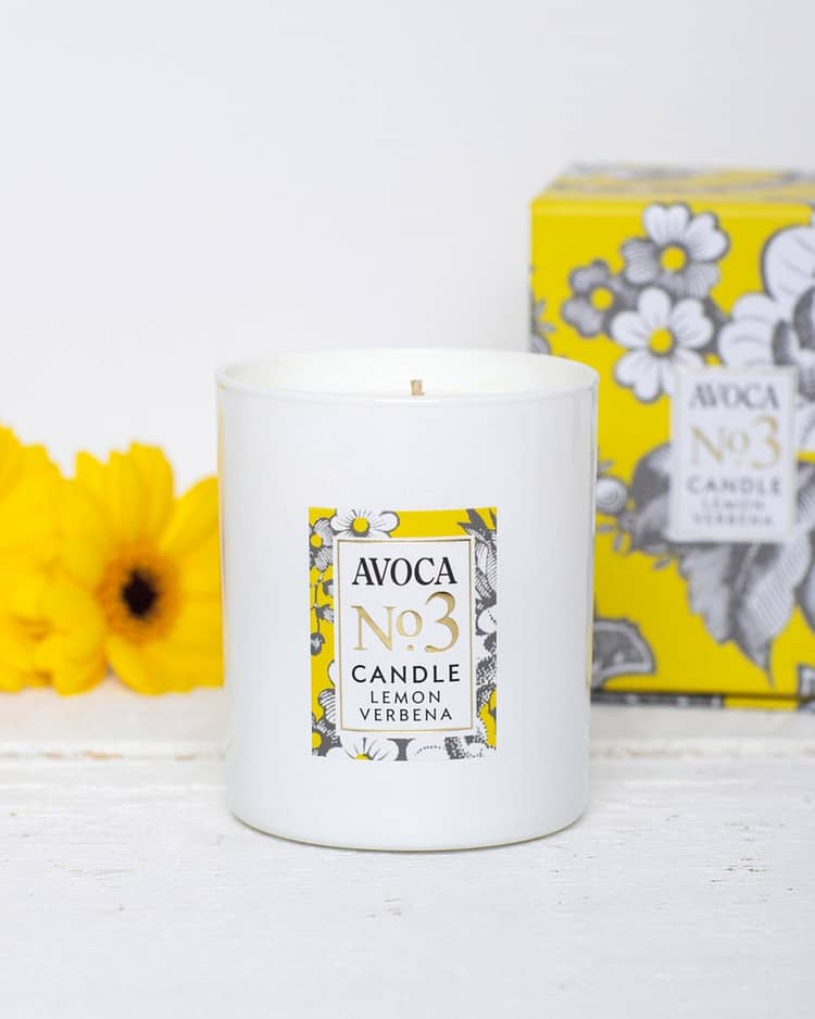 Avoca Candle lemon verbena in a white container and yellow nametag with flowers on the front of container, Yellow flowers and the packaging box for the candle are in the background against a white backdrop wall