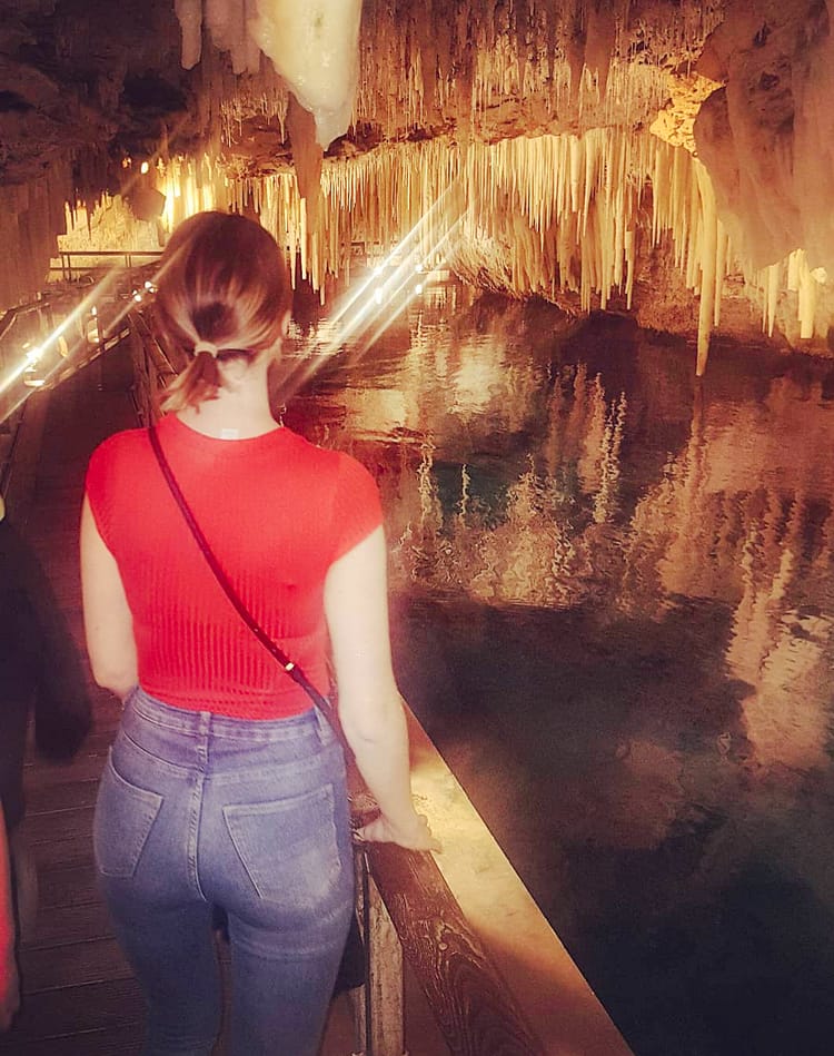 Girl in red top and jeans, with back to camera, looking over water and hanging stalactites and stalagmites in Crystal Cave, Bermuda.