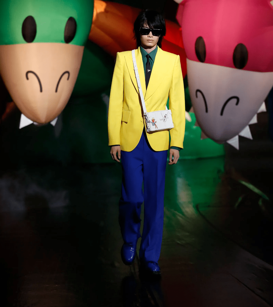 Male model walks a show in a yellow jacket and blue trousers with sunglasses on. Two balloon animal shapes are in the background.